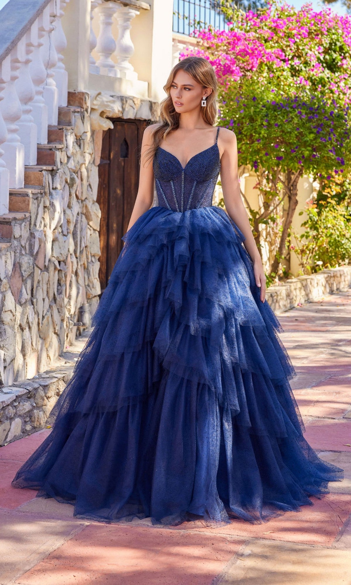 A woman stands outdoors in sunlight wearing the Long Prom Dress: Juliet JT2611N, a dark blue strapless gown with a fitted bodice, surrounded by stone walls and blooming flowers.
